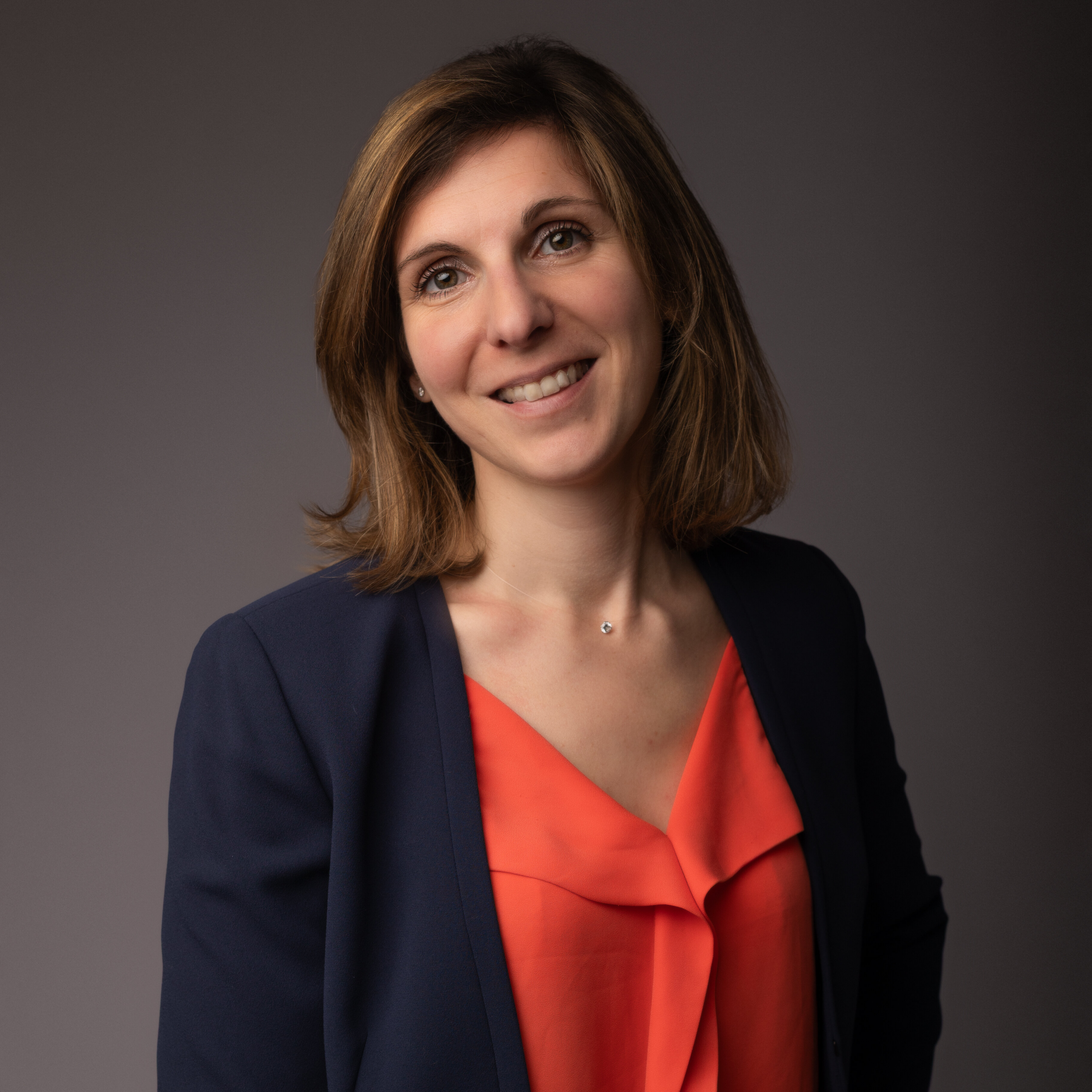 Smiling woman with shoulder-length brown hair, wearing a navy blazer and orange blouse, against a gray backdrop.