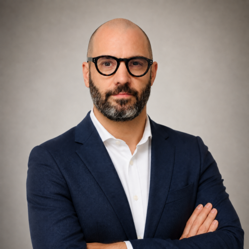 Professional headshot of a bald man with a beard, wearing round black glasses and a navy suit, arms crossed against a neutral backdrop.