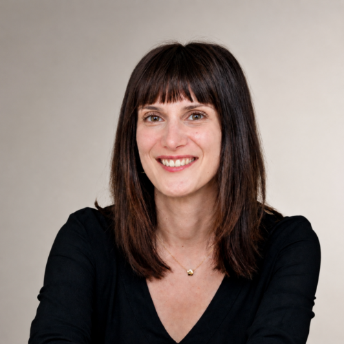 Smiling woman with shoulder-length dark hair and bangs wearing a black V-neck top and a small gold necklace, against a neutral backdrop.