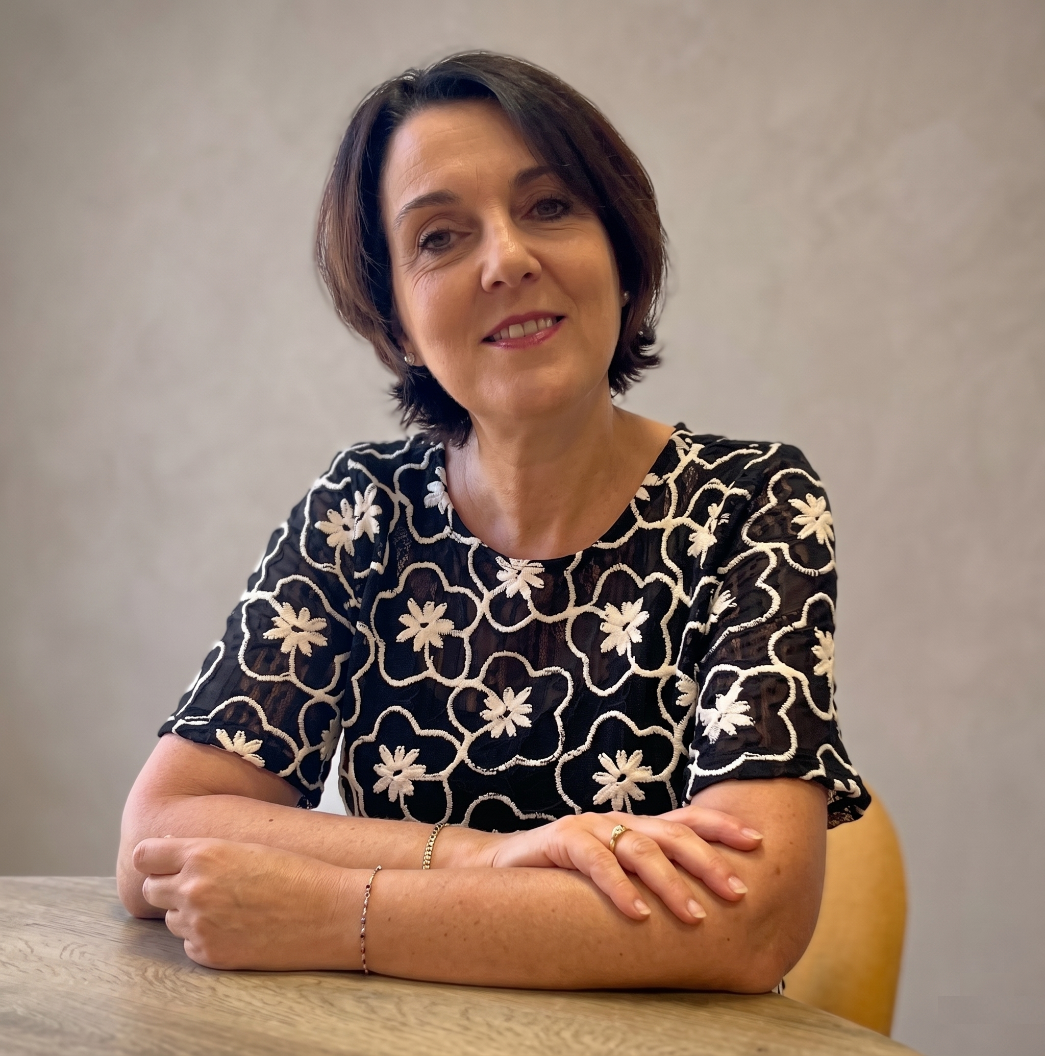 Smiling woman with short dark hair sitting at a wooden table with arms crossed, wearing a black dress with white floral embroidery.